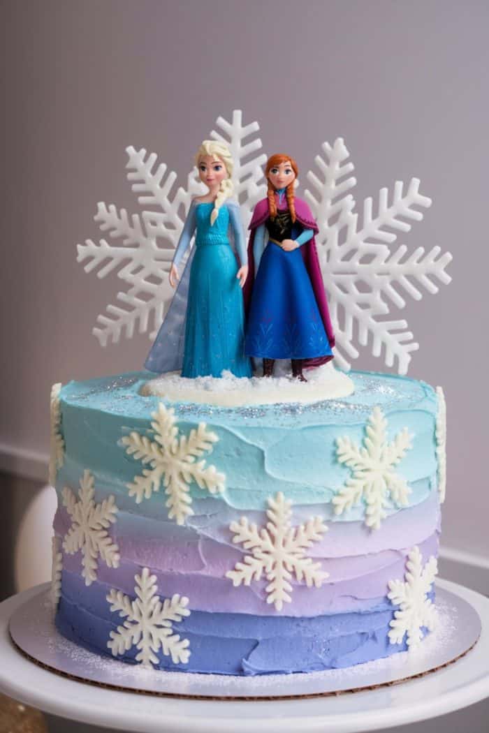 A layered cake with pastel blue and purple frosting, decorated with edible snowflakes. Topped with two figures resembling characters from a popular animated movie. The theme evokes a wintery, magical feel.