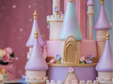 A whimsical, multi-tiered cake designed as a pastel-colored fairytale castle with pink, lavender, and mint towers. It features intricate details like golden doors, turrets, and floral decorations. The backdrop is shimmering pink.