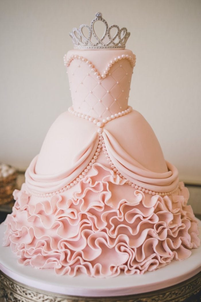 A pink princess dress cake adorned with a quilted bodice, pearl-like decorations, and ruffled skirt layers. Topped with a silver tiara, the cake resembles a ball gown, set on a white platter.