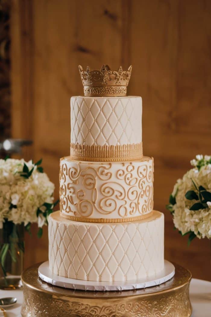 Three-tiered white wedding cake with gold ornate patterns and a gold crown topper. The tiers are intricately decorated with swirls and diamond patterns. The cake sits on a decorative gold stand, surrounded by vases of white flowers.
