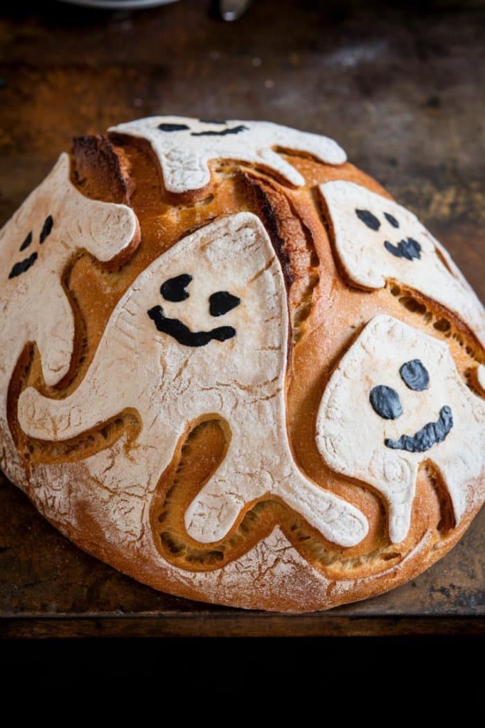 A round loaf of bread decorated with playful, ghost-shaped designs on the crust. The ghosts have smiling faces drawn in black, adding a whimsical touch to the golden-brown surface. The bread sits on a rustic wooden surface.