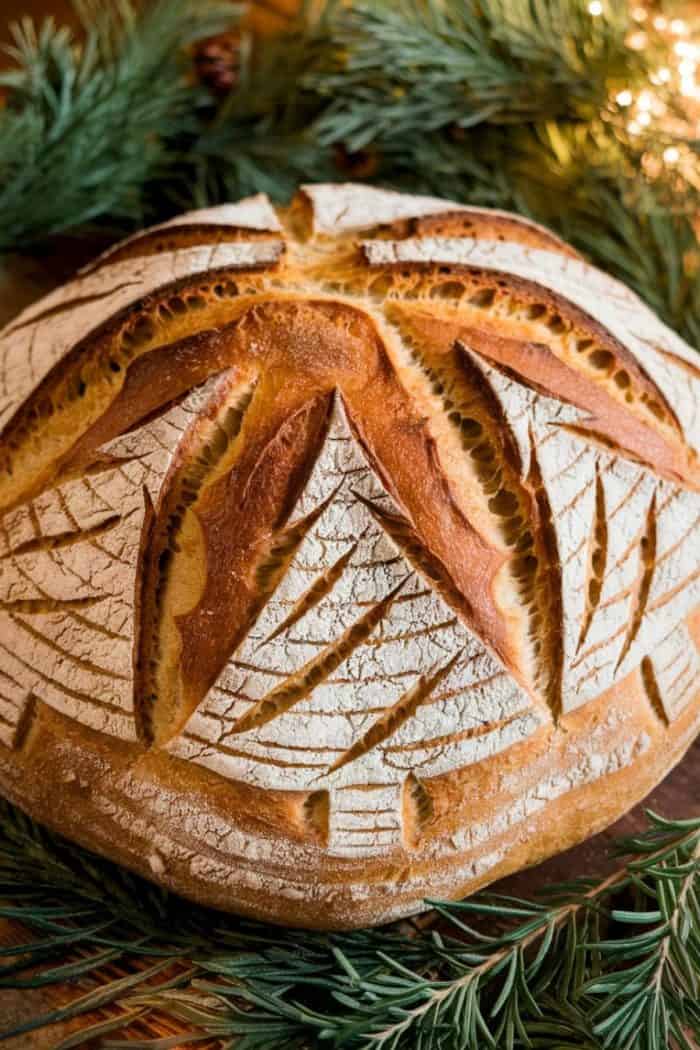 A round, artisan loaf of bread with intricate scoring patterns on top, resembling star shapes. The bread's crust is golden-brown, and it rests on a bed of green pine branches, illuminated softly from the side.