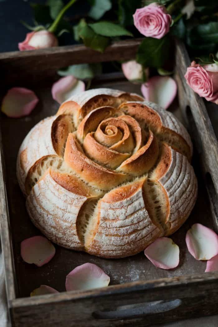 A beautifully crafted round loaf of bread in the shape of a rose, placed in a wooden tray. The loaf has a golden-brown crust with intricate scoring. Pink roses and petals surround the bread, creating an elegant presentation.