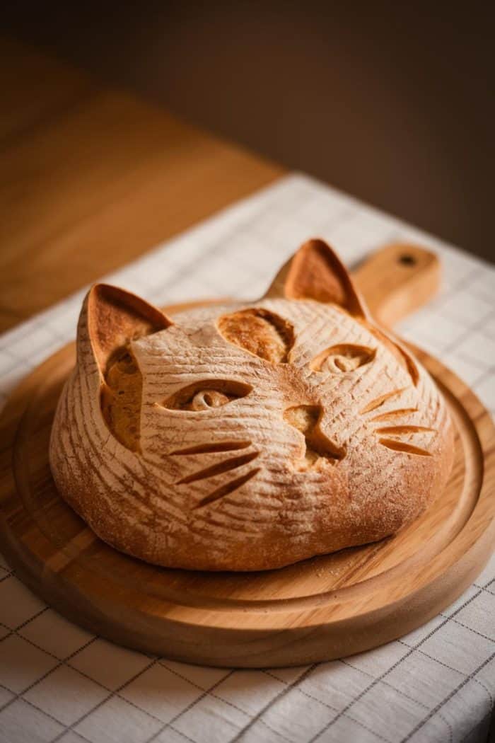 A loaf of bread shaped and decorated to resemble a cat's face, complete with ears and whiskers, is placed on a round wooden cutting board. The bread is on a checkered cloth with a warm light illuminating the scene.