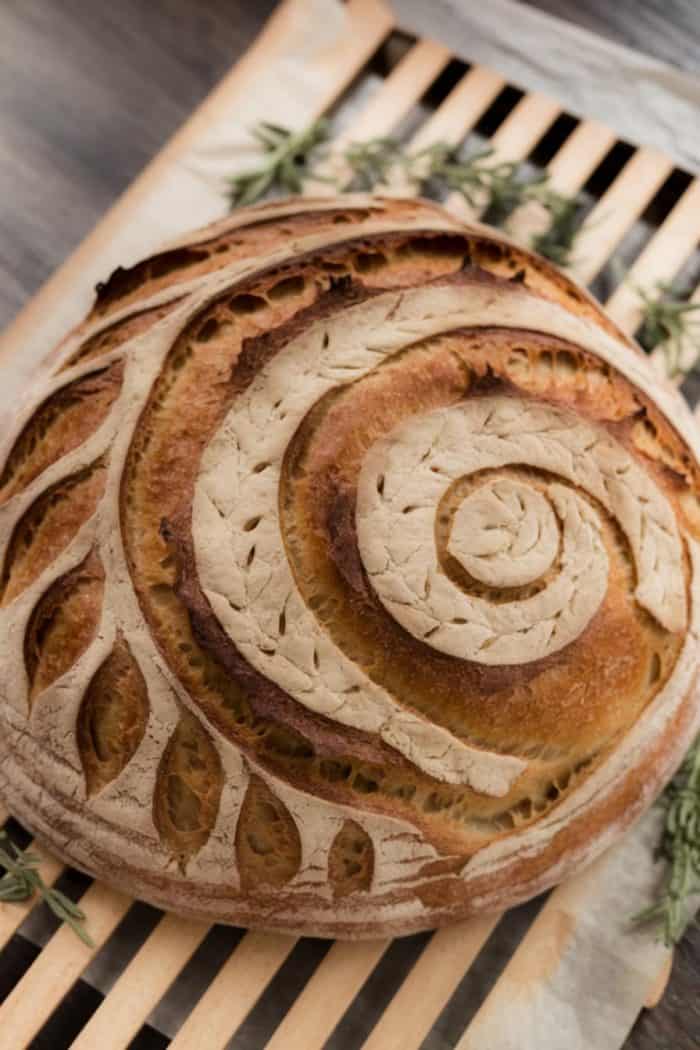 Round artisan loaf with a spiral and leaf pattern etched on the crust, resting on parchment paper atop a wooden slatted board. Sprigs of fresh herbs are nestled around the bread.