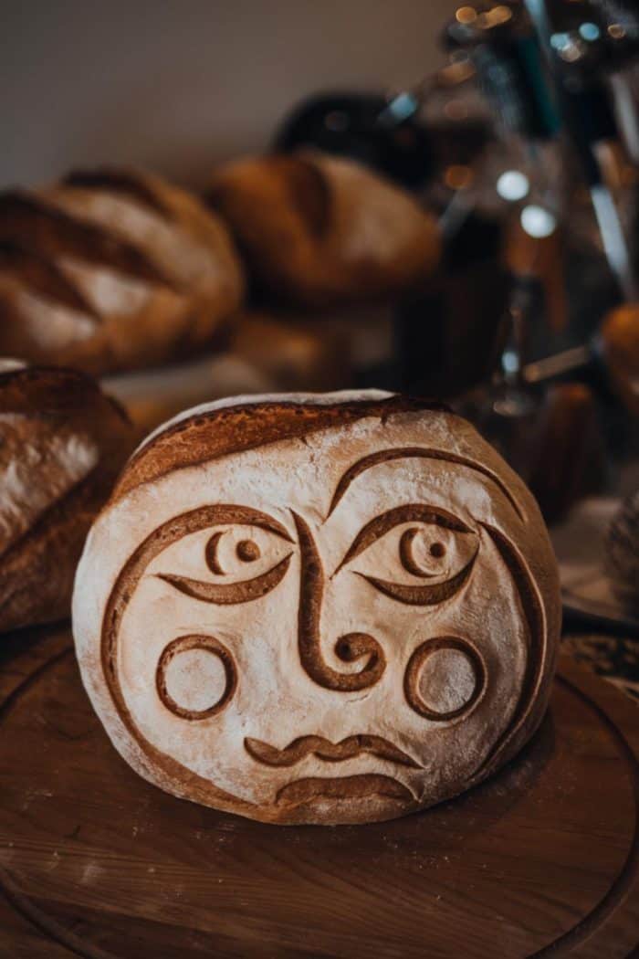 A round loaf of bread with a carved face featuring two eyes, a nose, a mouth, and two circular cheeks. Other loaves of bread are blurred in the background.