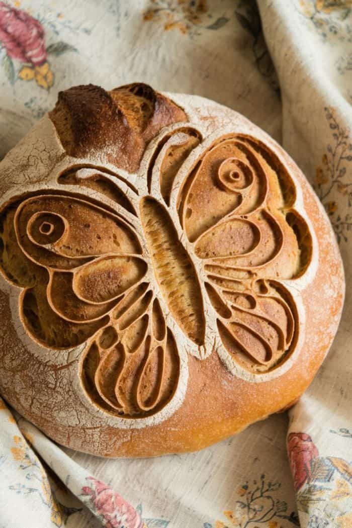 Round loaf of bread with an intricate butterfly design scored on top, resting on a floral-patterned fabric.