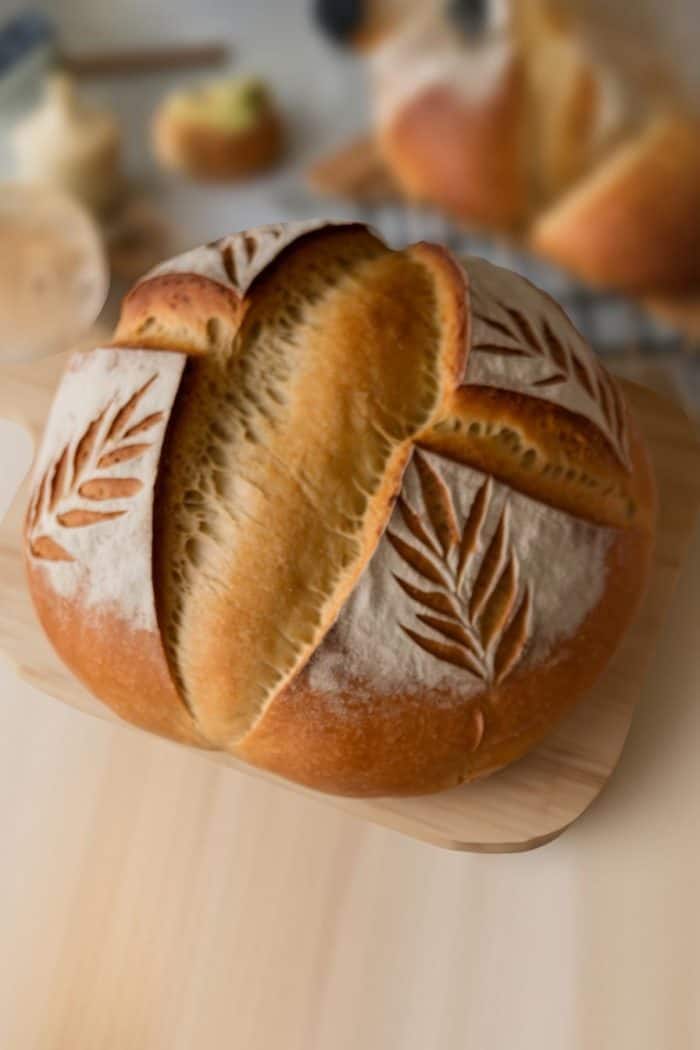 Round loaf of artisan bread with a decorative pattern of leaves scored on top. The crust is golden brown, and the bread is placed on a wooden board. Other loaves and ingredients are blurred in the background.