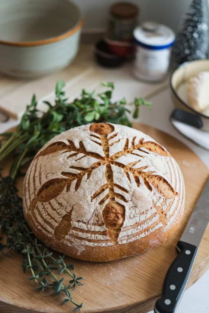 A round loaf of artisan bread with decorative scoring sits on a wooden cutting board. Fresh herbs are placed nearby, along with a serrated knife. In the background, there are kitchen items and a butter dish.