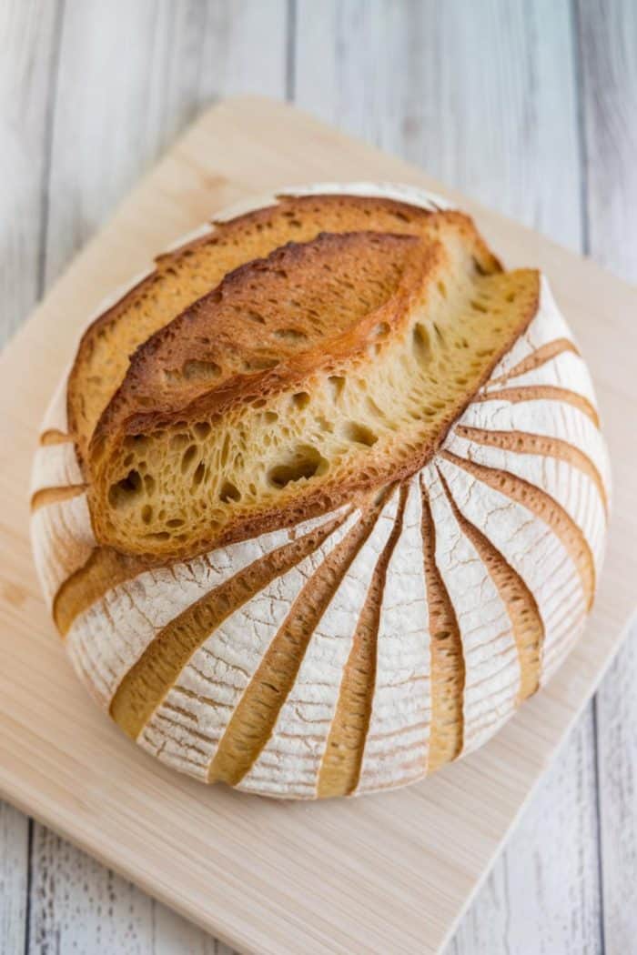 Round artisan bread with a decorative scoring pattern and a golden crust sits on a wooden cutting board. The bread's surface features a fan-like design, and it has a rustic, appealing appearance.