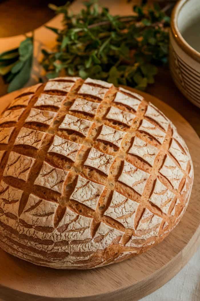 Round loaf of crusty bread with a crisscross pattern on top, resting on a wooden board. Fresh herbs and a bowl are in the background, creating a warm and rustic kitchen setting.