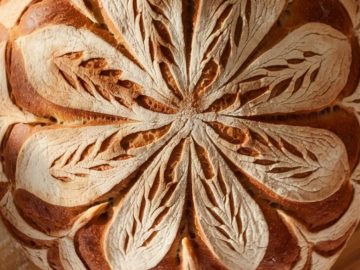 A round loaf of artisan bread with intricate leaf patterns scored into the crust, showcasing a golden-brown color. The bread rests on a wooden surface, highlighting the detailed design and rustic appearance.