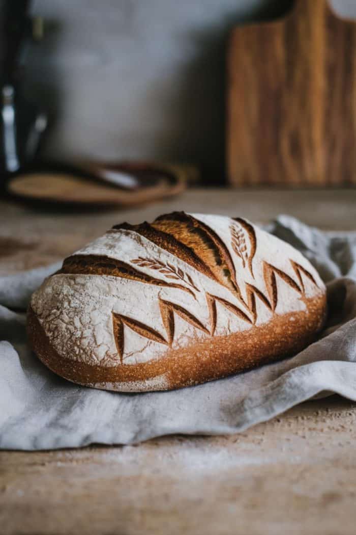 A loaf of artisan bread with decorative scoring on the surface sits on a cloth. The bread has a golden crust and appears freshly baked, resting on a rustic wooden surface with a cutting board in the background.