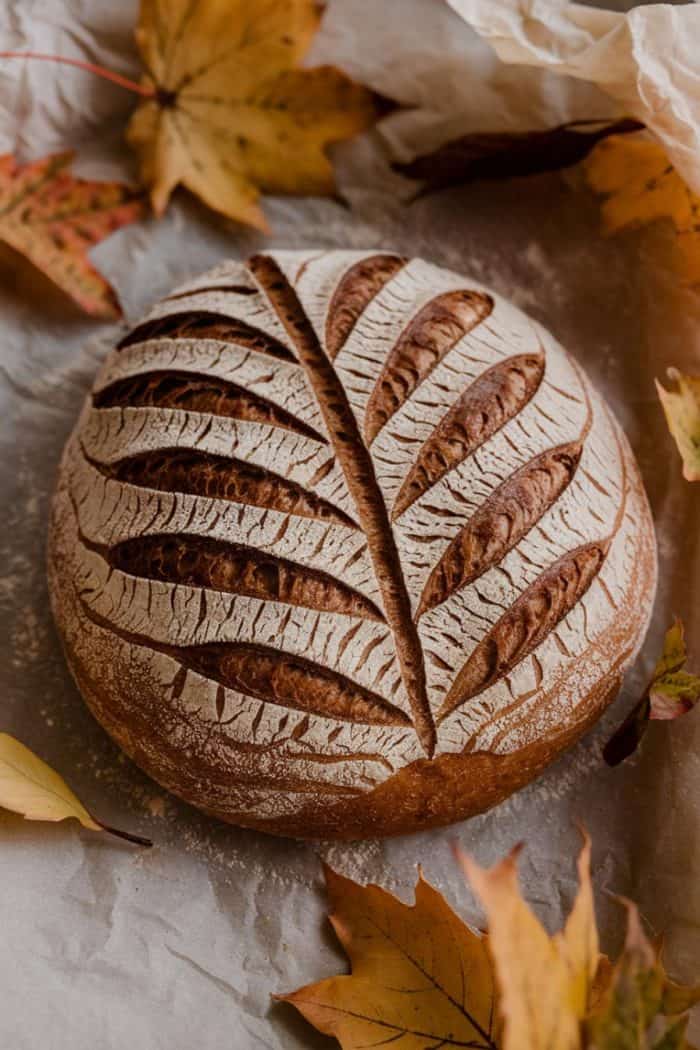 A round loaf of bread with intricate leaf designs scored on the crust, resting on parchment paper. Surrounding the bread are various autumn leaves in shades of yellow and orange.