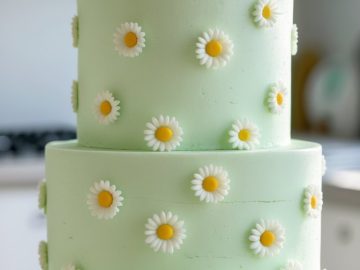 A two-tiered light green cake decorated with evenly spaced white and yellow daisy flowers sits on a white cake stand in a bright kitchen.