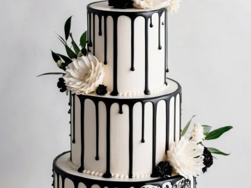 A three-tier white cake with black drip decoration and intricate black floral patterns. It is adorned with large white and black flowers and green leaves on each tier, placed on a white cake stand against a light background.
