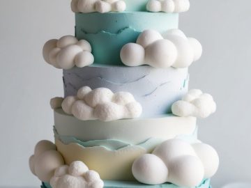 A tiered cake designed to look like a stack of clouds. Each layer is a light blue gradient with fluffy, white cloud-shaped meringues. The cake is displayed on a white surface against a plain background.