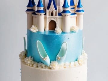 A two-tiered cake decorated with blue and white frosting, featuring a castle topper and two light blue fondant slippers. The bottom tier has a ruffled blue design, and the cake is surrounded by blue spoon-like decorations.
