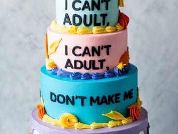 A four-tiered colorful cake with a graduation cap topper. The tiers are decorated with frosting and say, “I CAN’T ADULT,” “I CAN’T ADULT,” and “DON’T MAKE ME.” The cake is on a white stand.