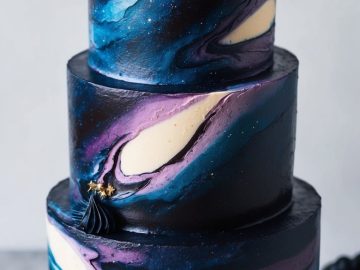 A three-tiered cake decorated with swirling galaxy patterns in dark blue, purple, and white, topped with blue meringue-like swirls and small gold star accents, displayed on a white cake stand.