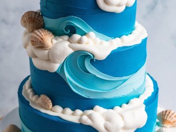 A two-tiered blue cake decorated with white frosting waves, seashells, and fluffy clouds, presented on a white cake stand. The design resembles an ocean scene with shells around the base and tiers.