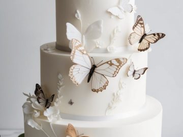 A three-tiered white cake decorated with realistic edible butterflies in shades of brown and cream, along with delicate white sugar leaves, sits on a marble surface with gold utensils and a sprig of greenery nearby.