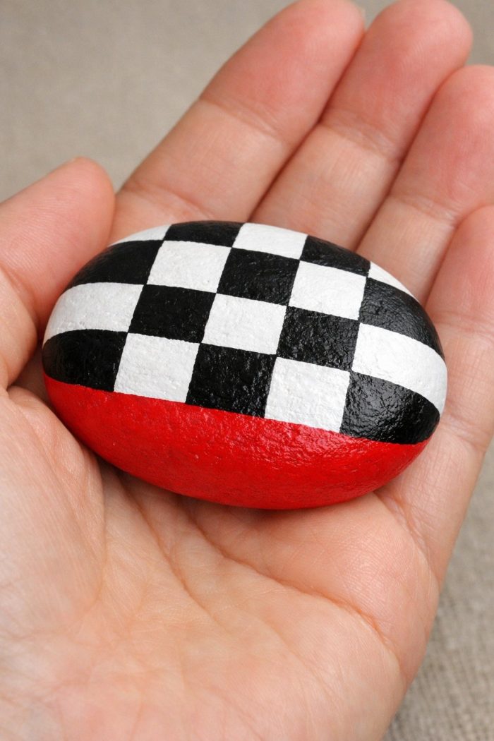 A hand holds a smooth oval stone painted with a black and white checkerboard pattern on top and a solid red section along the bottom.