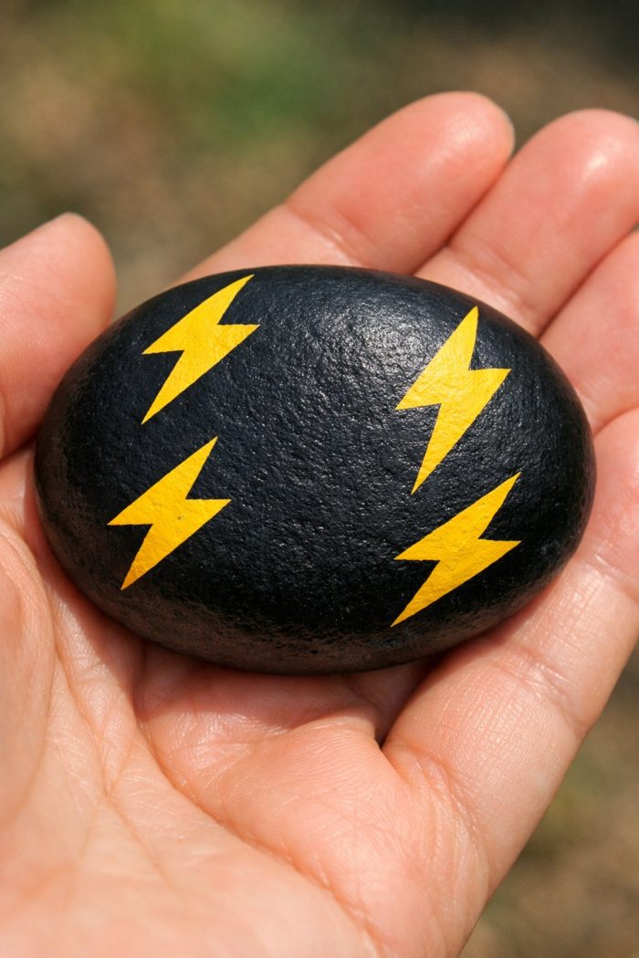 A hand holds a smooth, black stone painted with five yellow lightning bolt symbols on its surface. The background is blurred greenery.