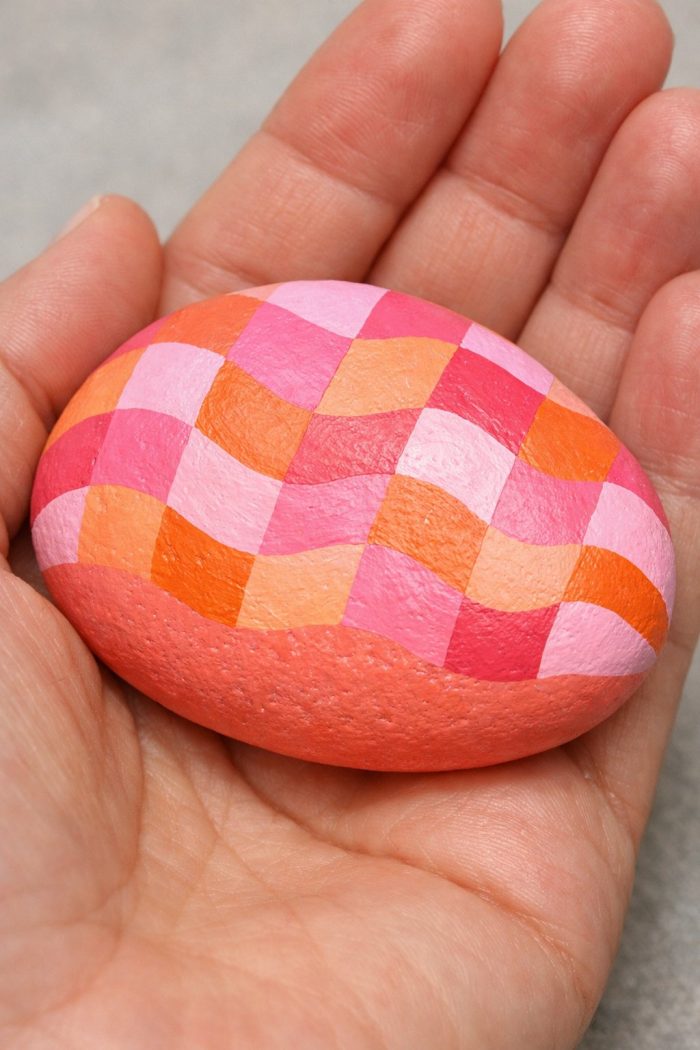 A hand holds a smooth oval stone painted with a checkered wavy pattern in shades of pink, orange, and coral.