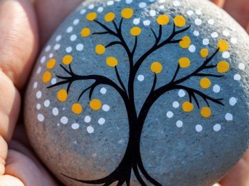 A hand holds a smooth gray stone painted with a black tree, featuring branches decorated with yellow and white dots resembling leaves or blossoms.