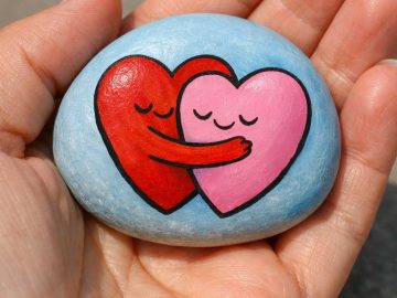 A hand holds a smooth stone painted with a red heart and a pink heart, both with closed eyes and smiling, as the red heart hugs the pink heart on a light blue background.