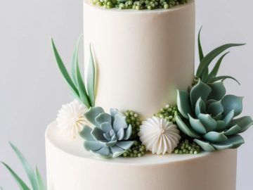 A two-tiered white cake decorated with realistic green succulent plants and small white flowers made from icing, displayed on a white cake stand against a plain background.