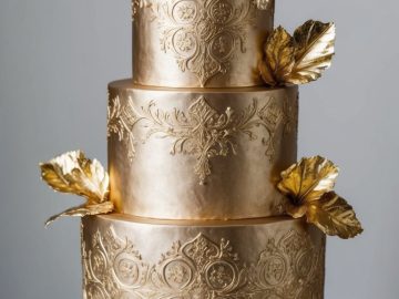 A three-tiered gold cake with intricate embossed patterns and decorated with metallic gold leaves, displayed on a white cake stand with a gold base. Plates and a fork are set nearby.