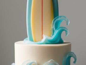 A round cake decorated with blue icing waves and topped with a large, upright surfboard made of fondant, creating a beach or surfing theme.