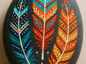 A smooth black stone painted with three colorful feathers—blue, red, and orange—each with intricate patterns and white highlights, on a light beige background.