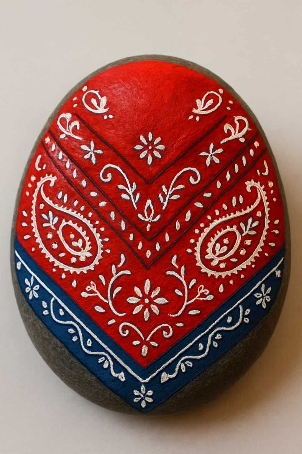 Close-up image of a smooth river rock painted with a colorful cowboy bandana design featuring paisley patterns.