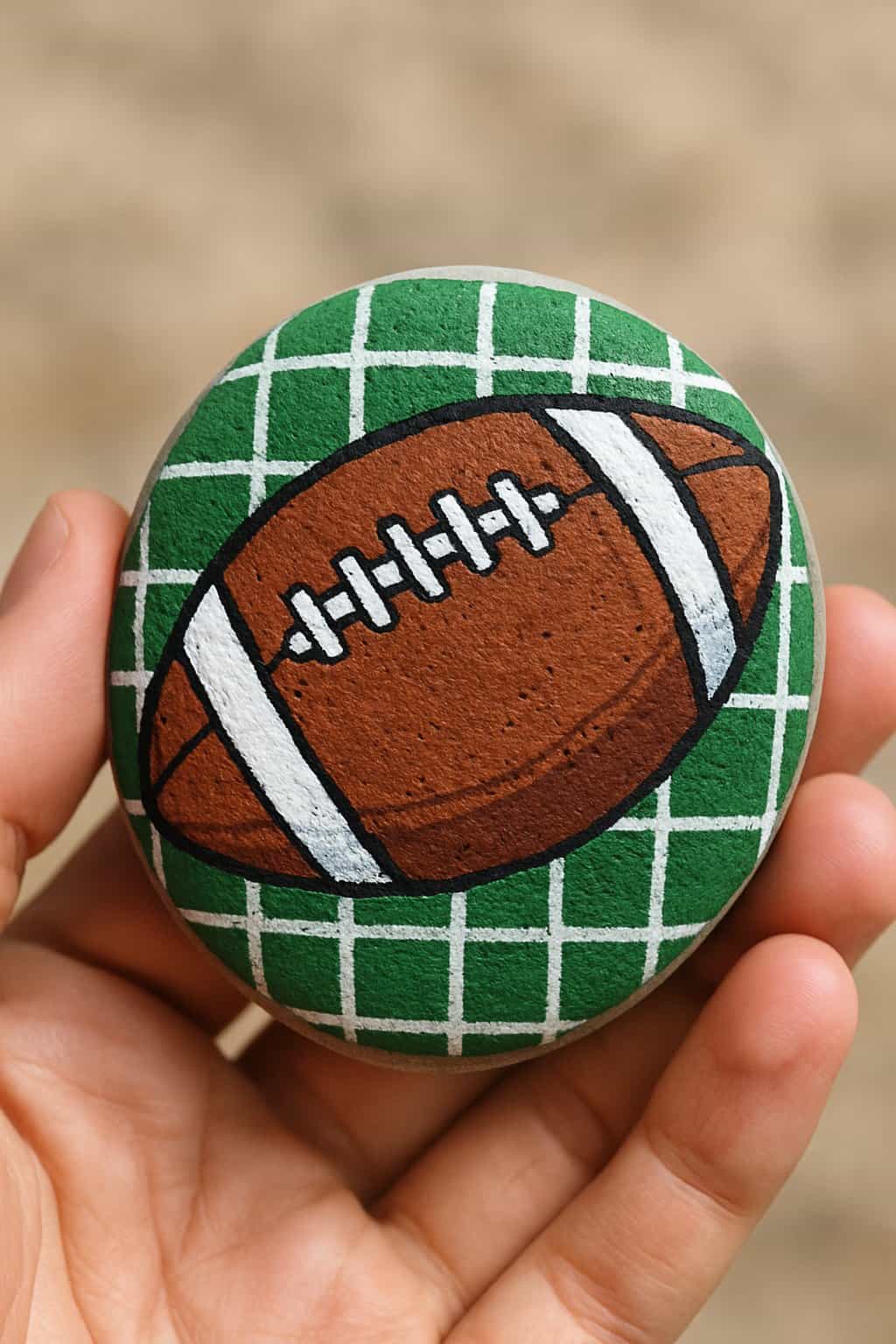 A hand holding a smooth river rock painted with a football and gridiron pattern.