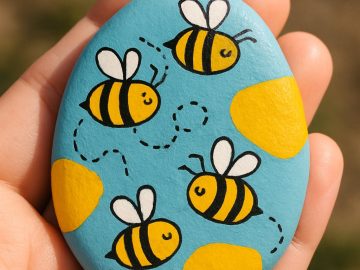 A hand holds a blue painted stone featuring three cartoon bees with dotted flight paths and two yellow spots, set against a blurred outdoor background.
