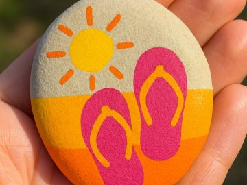 A hand holds a smooth stone painted with a yellow sun and pink flip-flops on an orange and yellow background, resembling a summer beach scene.