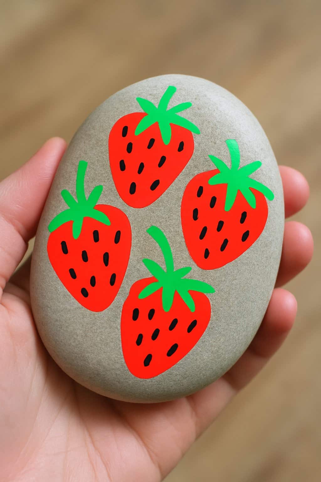 A hand holding a smooth rock painted with bright red and green strawberry designs.
