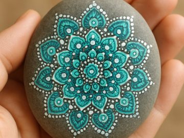 A hand holds a smooth stone painted with an intricate teal and white mandala pattern against a soft brown background.