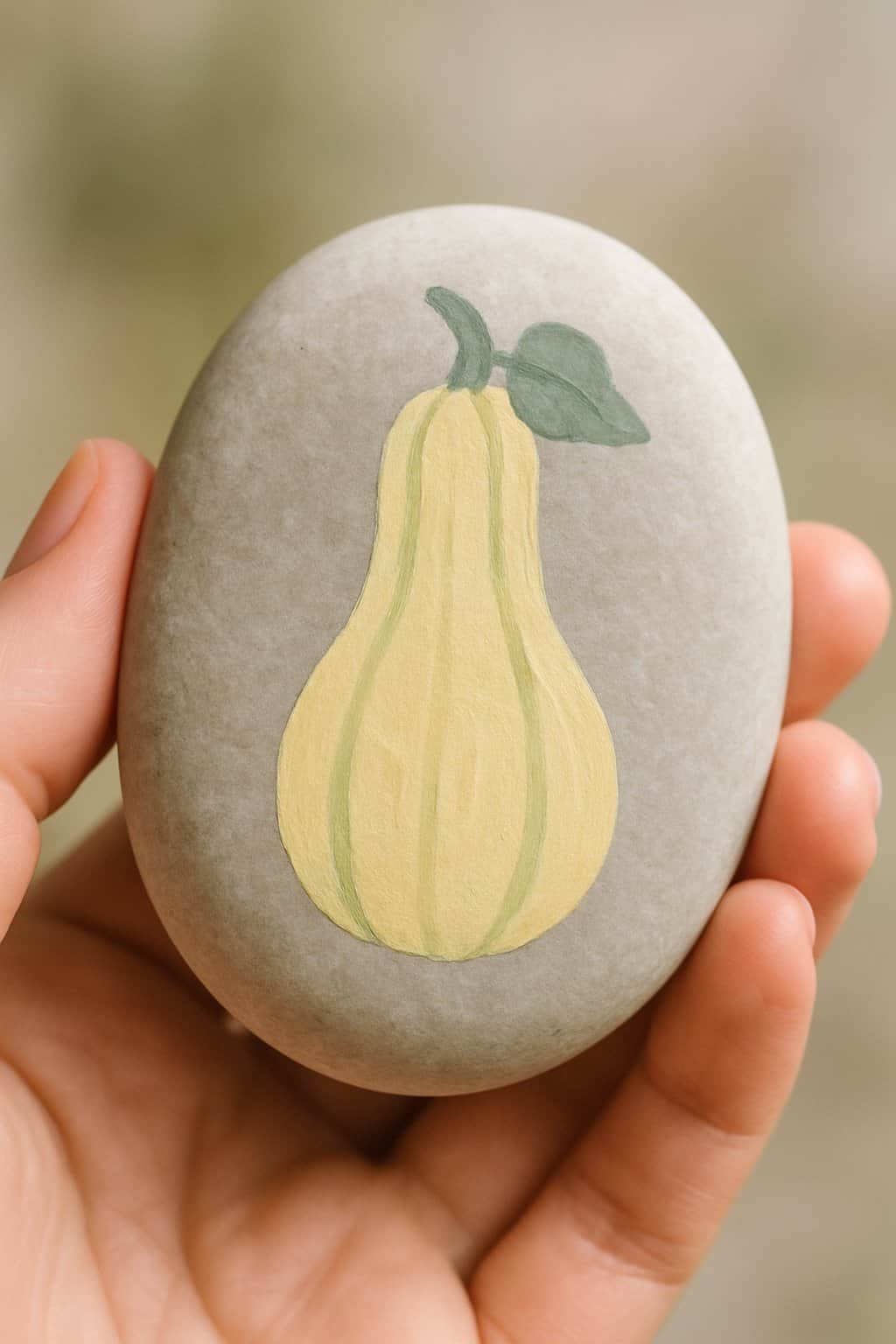 A hand holding a smooth river rock painted with a squash design in pale yellow and soft green.