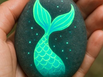 A hand holds a smooth stone painted with a glowing green mermaid tail design, featuring scales and surrounded by small dots, against a blurred outdoor background.