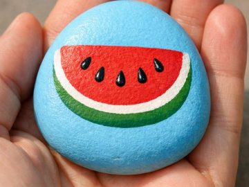 A hand holds a smooth, round stone painted blue with a colorful image of a watermelon slice, featuring red flesh, black seeds, and a green and white rind.