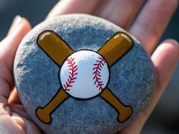 A hand holds a smooth gray stone painted with two crossed baseball bats and a white baseball with red stitching in the center.