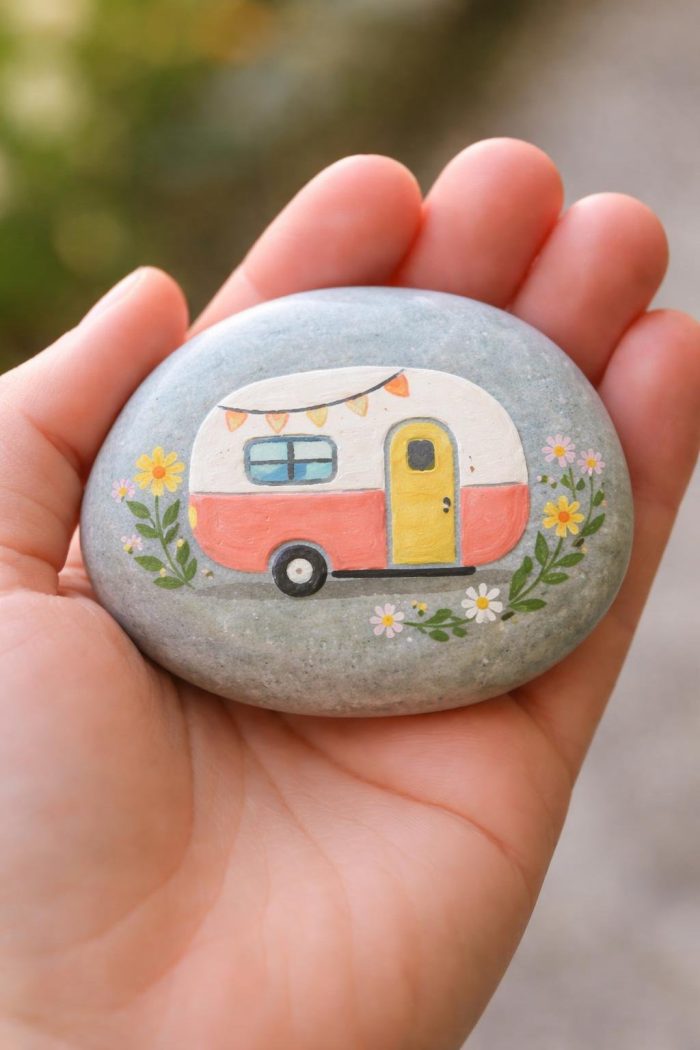 A hand holds a smooth stone painted with a cute camper van, featuring a yellow door, bunting, and floral decorations on either side. The background is blurred.