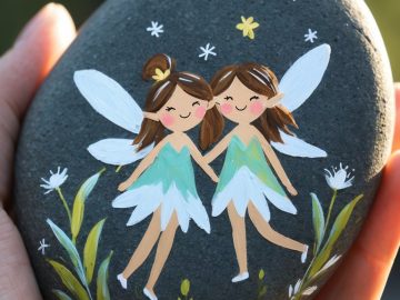 A hand holds a smooth dark stone painted with two smiling fairies in green and white dresses, white wings, and surrounded by flowers and stars.