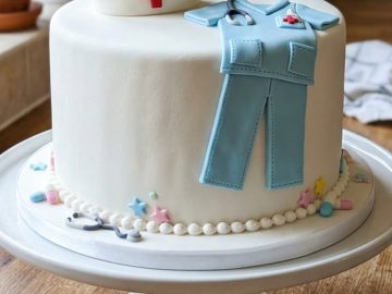 A white cake decorated with a nurse's cap, blue scrubs, and medical-themed fondant details sits on a white cake stand in a kitchen.