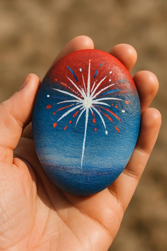 A hand holds a smooth stone painted red and blue, featuring a white firework with red and blue accents bursting in the center. The background is out of focus.