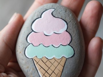 A hand holds a smooth gray stone with a colorful ice cream cone, featuring three scoops, painted on its surface. The ice cream scoops are pink, white, and light green.